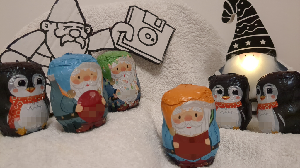 Mehrere Scholaden-Wichtel und -Pinguine auf einer weißen Decke blicken in die Kamera. Im Hintergrund weist ein großer Papp-Datenzwerg den Weg mit einer Diskette und ein weiterer Wichtel leuchtet mit seiner weißen Nase.